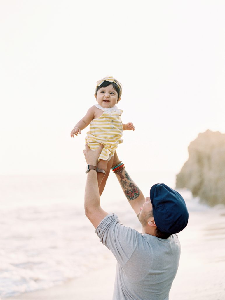 A dad holds his smiling baby daughter up in the air during their Malibu family portrait session with photographer Daniele Rose