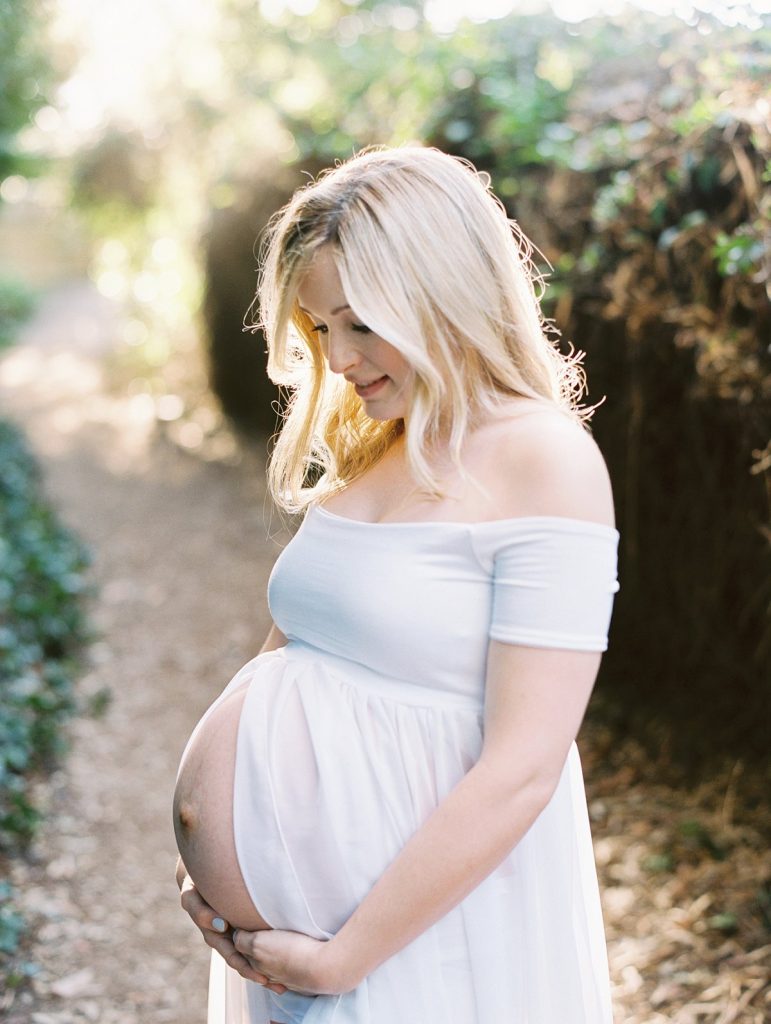 A pregnant woman in a white dress looks down at her belly in this maternity portrait