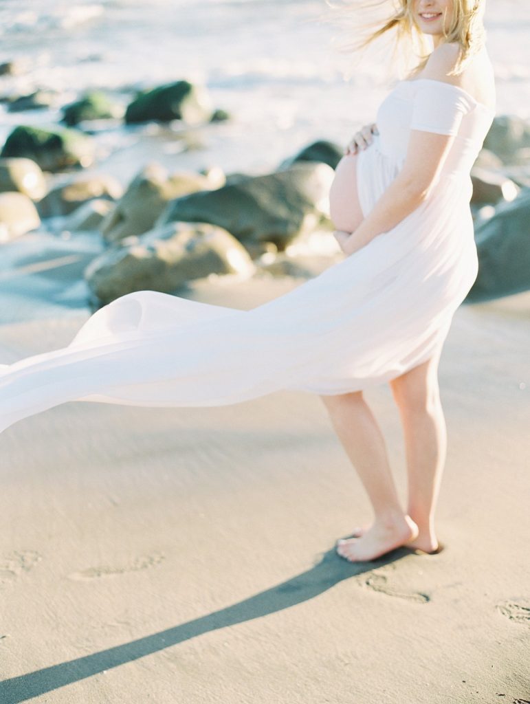 A pregnant woman stands on the sand at a beach in Santa Barbara while her white dress blows in the wind