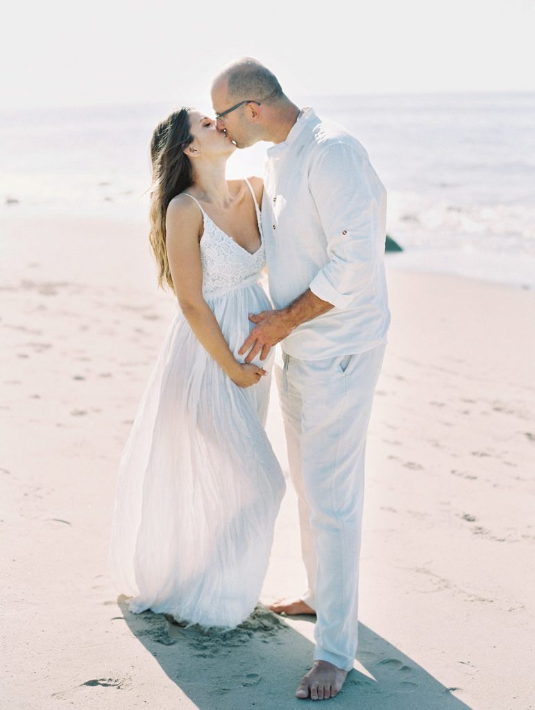A husband and wife wearing white kiss on the beach during their Santa Barbara family portraits 