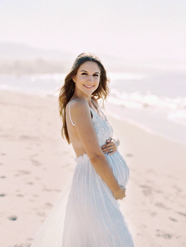 A pregnant woman wearing white smiles at the camera on the beach in Santa Barbara