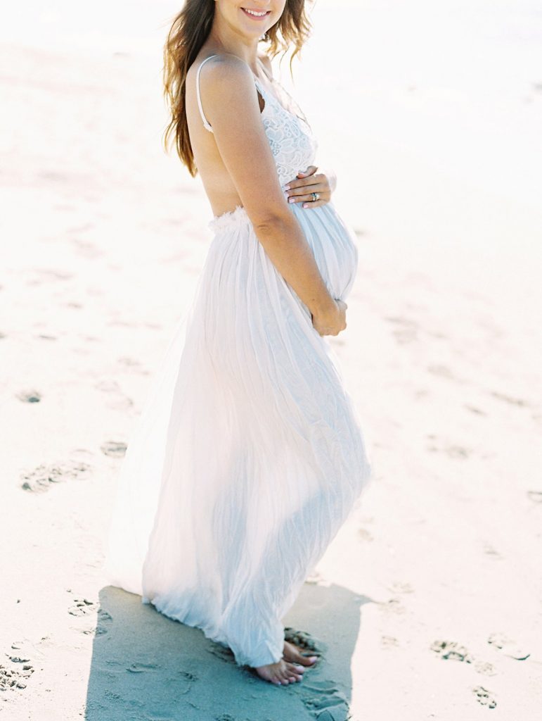 A maternity portrait of a mother to be in a white dress on the beach in Santa Barbara