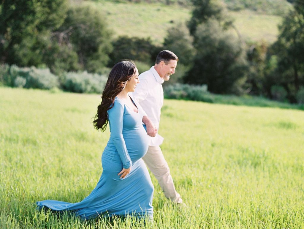 A couple walks hand in hand through a green field during their maternity portrait session in Thousand Oaks