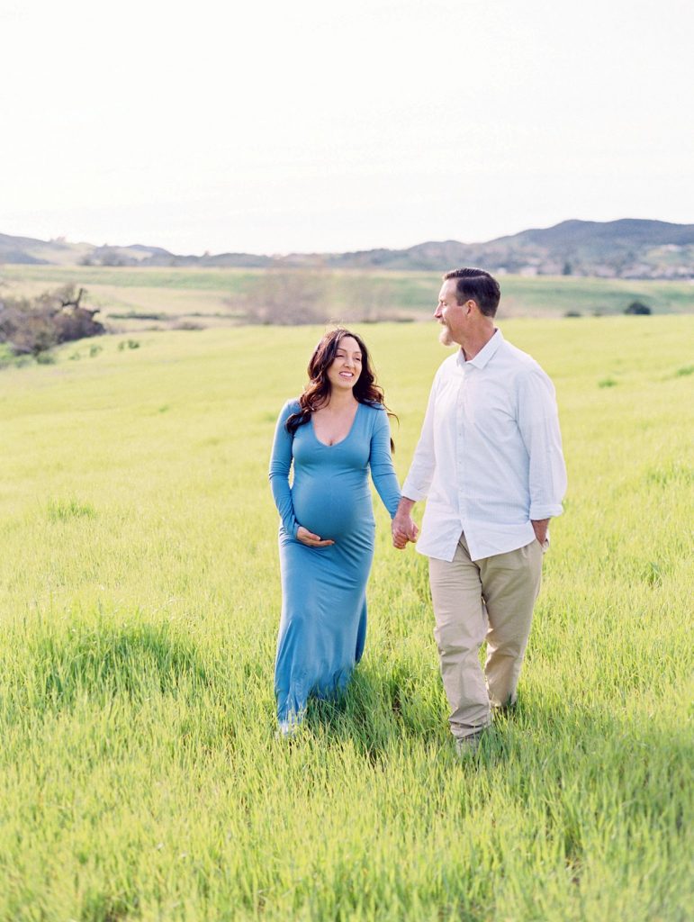 A husband and wife expecting their first child walk through a field of green grass in Thousand Oaks