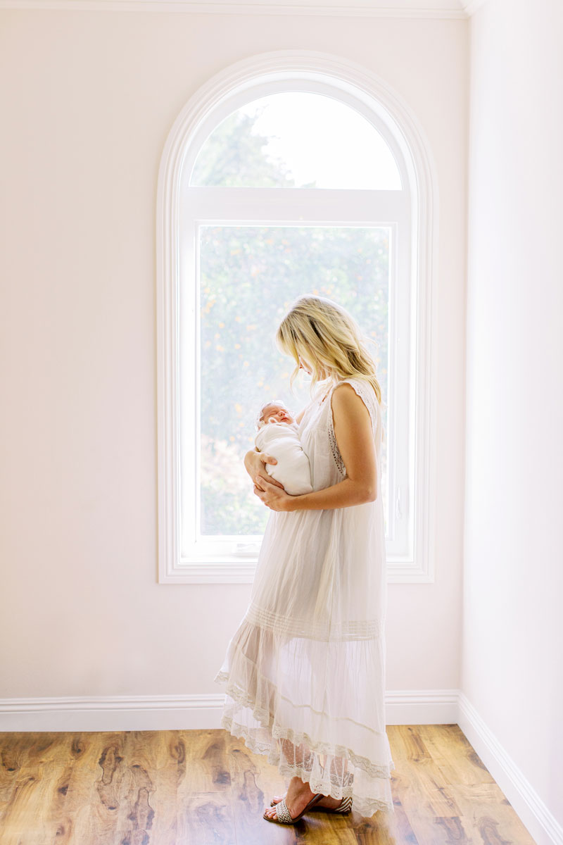 A mother wearing a white dress holds her newborn baby during her newborn photo session with Camarillo portrait studio photographer Daniele Rose
