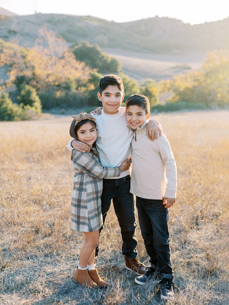 3 siblings hug in a golden field during their fall mini session with Ventura County family photographer Daniele Rose