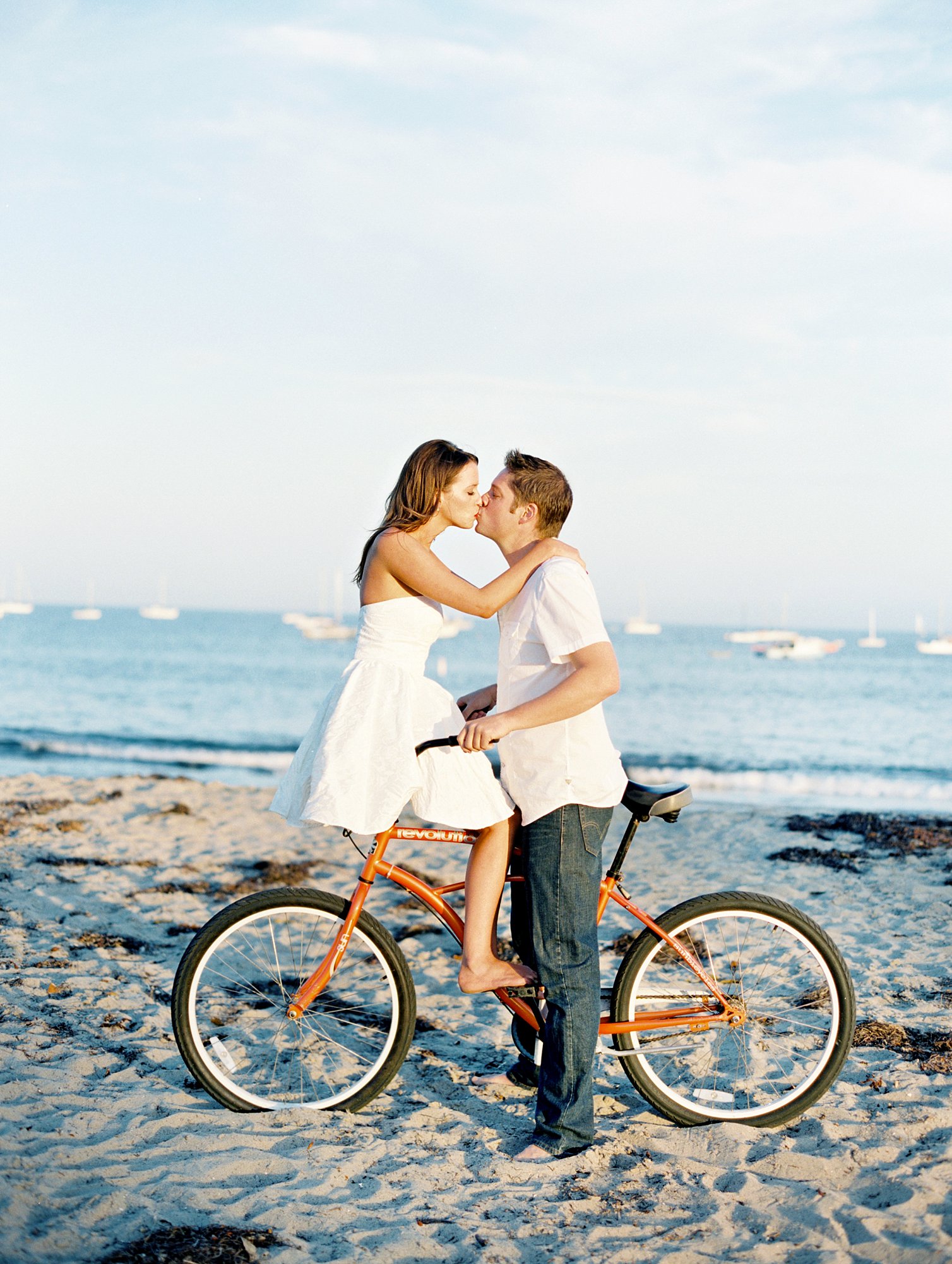 Image of a couple wearing white kissing while sitting on an orange bike in front of the ocean in Santa Barbara while on their babymoon.