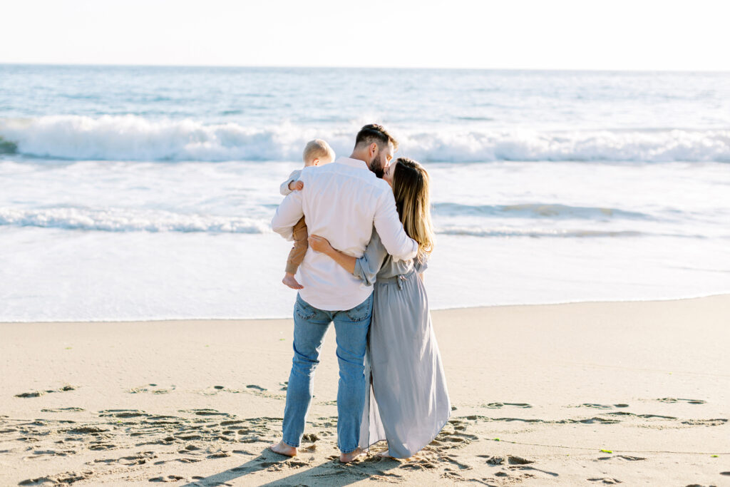 A mom and dad kiss on the beach while the dad hold their baby boy by Malibu photographer Daniele Rose
