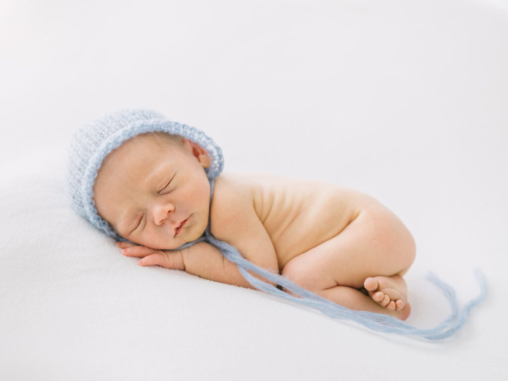 A posed naked baby wearing a blue knit cap lying on a white backdrop blanket.