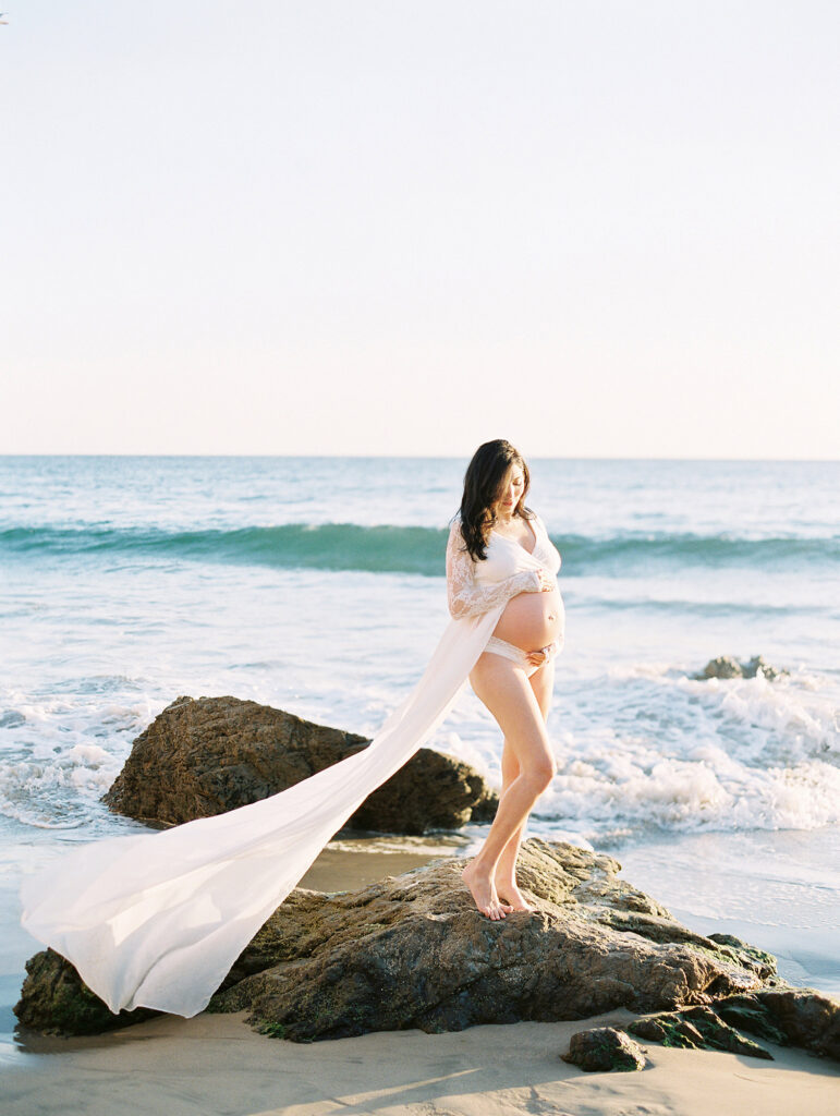 A mother-to-be wearing a white dress standing in the surf at El Matador Beach.