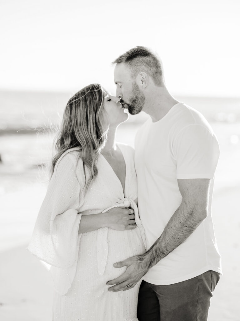 A couple wearing white kiss on the beach during their maternity photographer Los Angeles photo session.