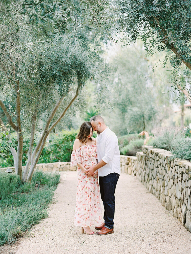 Ojai maternity photographer captures expecting couple embracing under olive trees on stone pathway