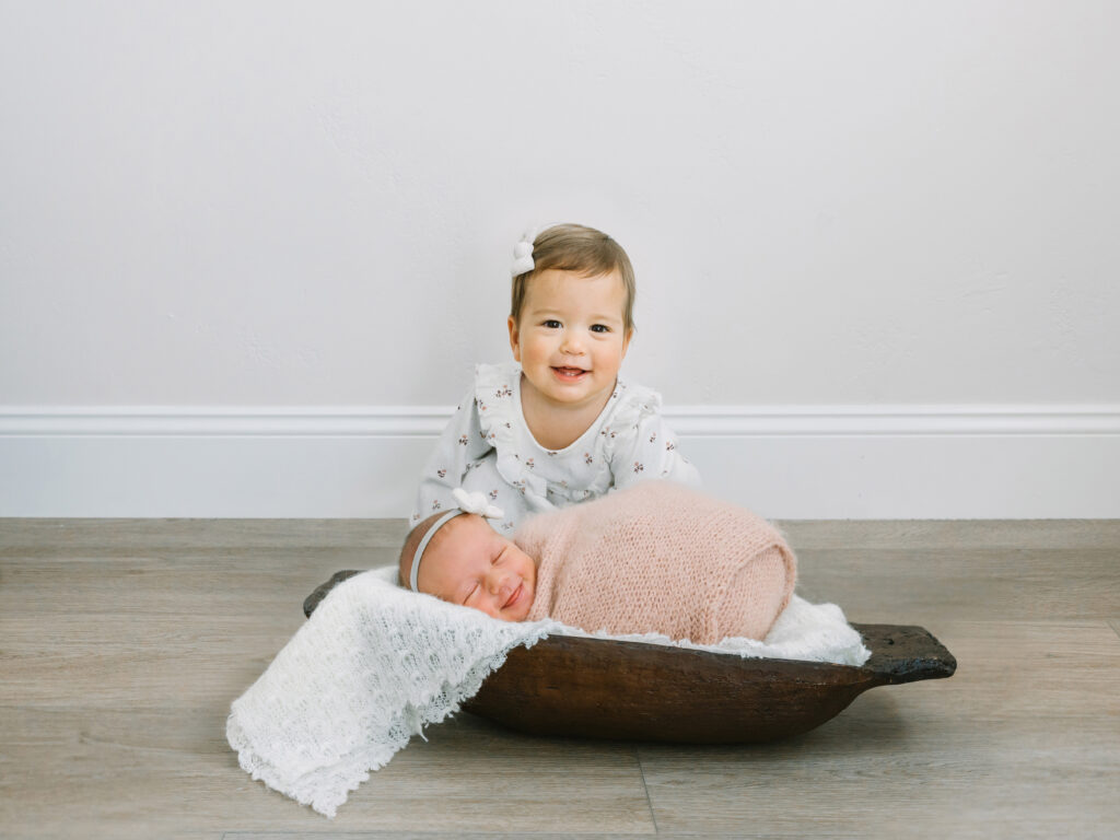 Older sibling sitting behind newborn baby in wooden bowl for safe and beautiful sibling portrait