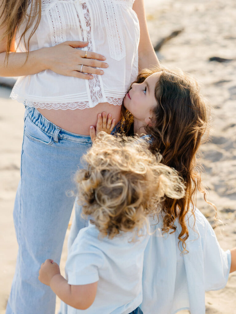 Young siblings cuddling Mom’s pregnant belly during family photos at the beach