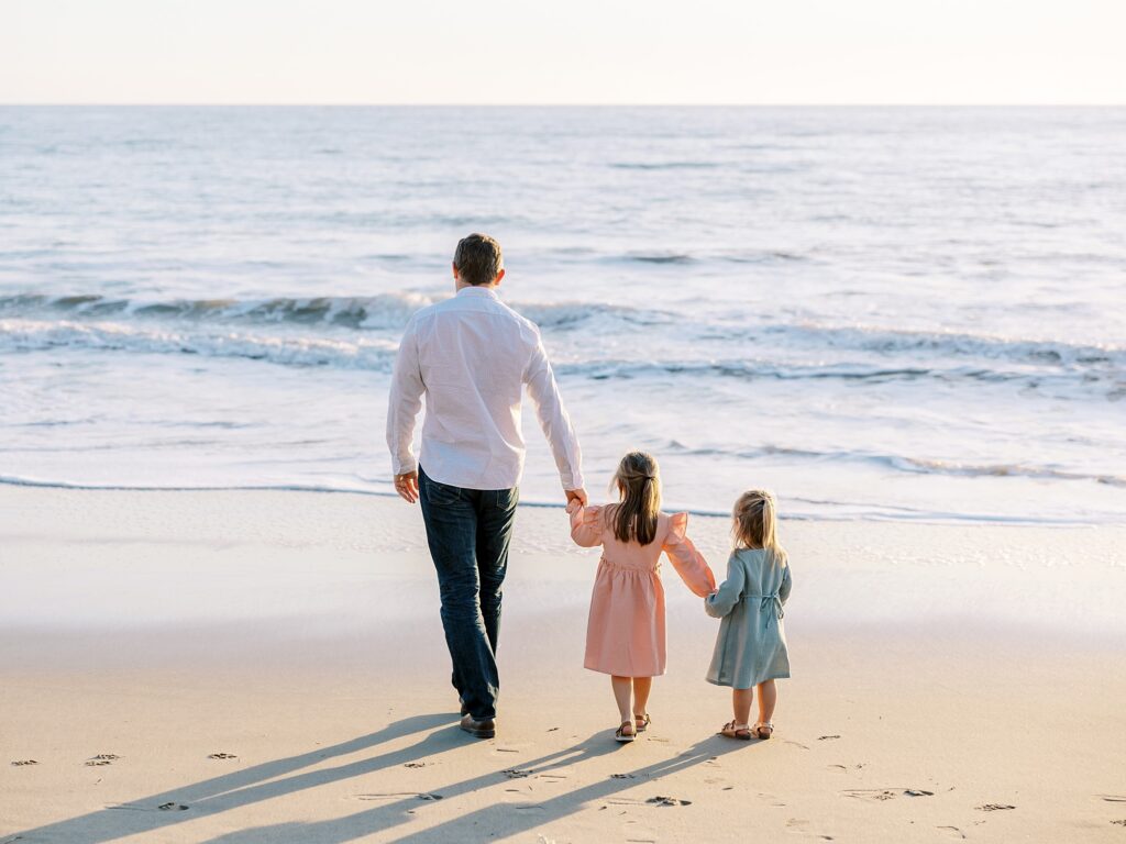 Father walking with two daughters toward the ocean waves during golden hour family photo session