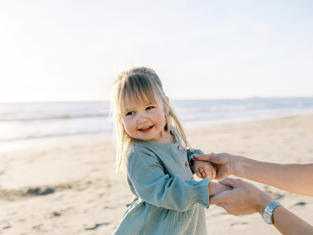 Toddler girl smiling and holding mother's hands on the beach, an example of the candid moments captured during full session when deciding between mini session vs full session