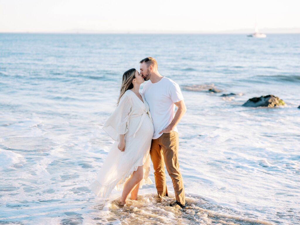 When to take maternity photos of an expecting couple kissing in the ocean at the beach