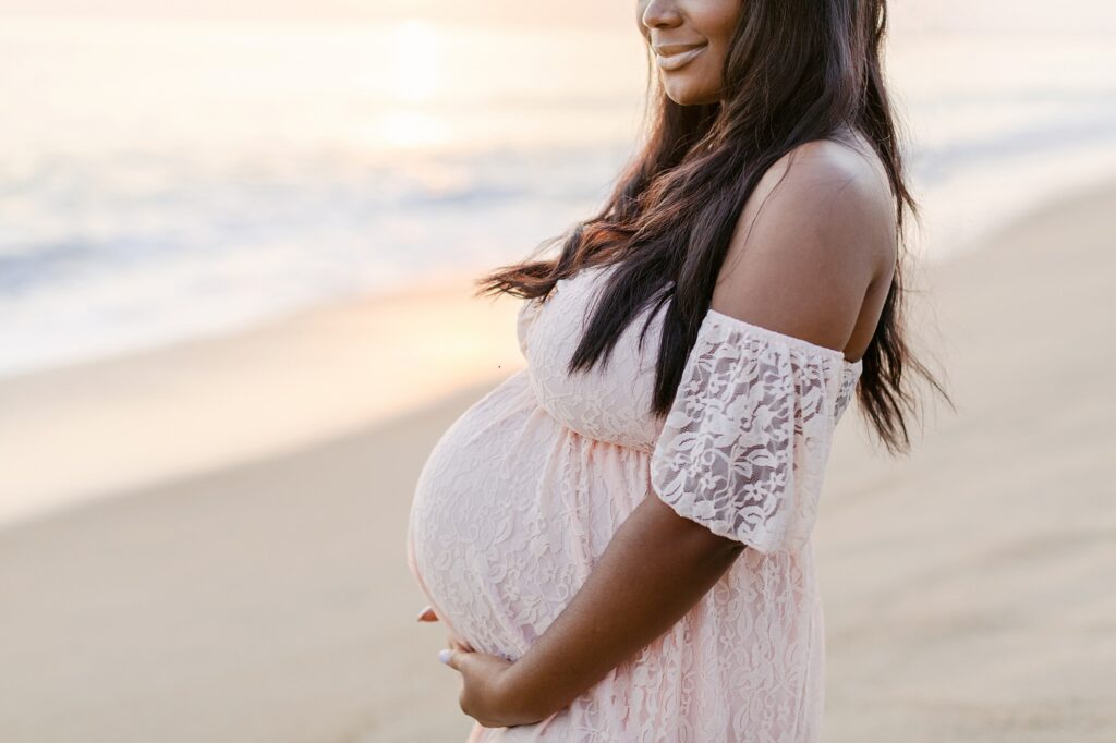 Expecting mother in a white lace dress showing her baby bump on the beach